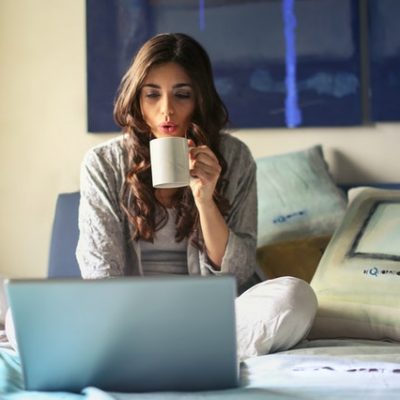 woman-in-grey-jacket-sits-on-bed-uses-grey-laptop-935743